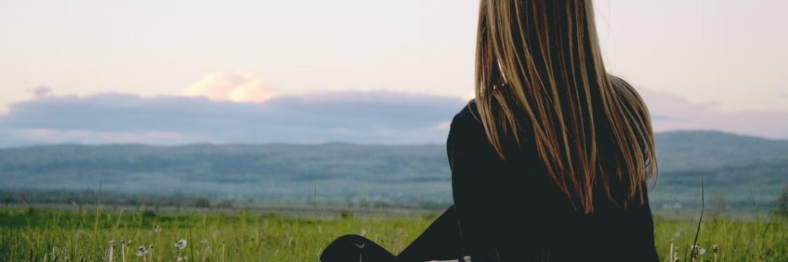 Woman enjoying peace in a lush green field with a scenic mountain view during sunset.