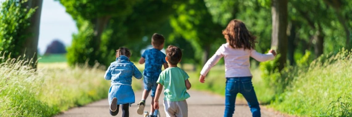 Four children on scooters and rollerblades moving along a tree-lined path in summer